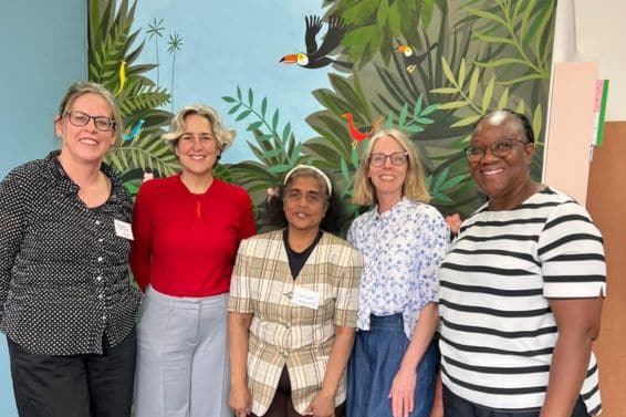 5 women standing in a line in front of a painted wall showing a scene of a bird flying through some trees
